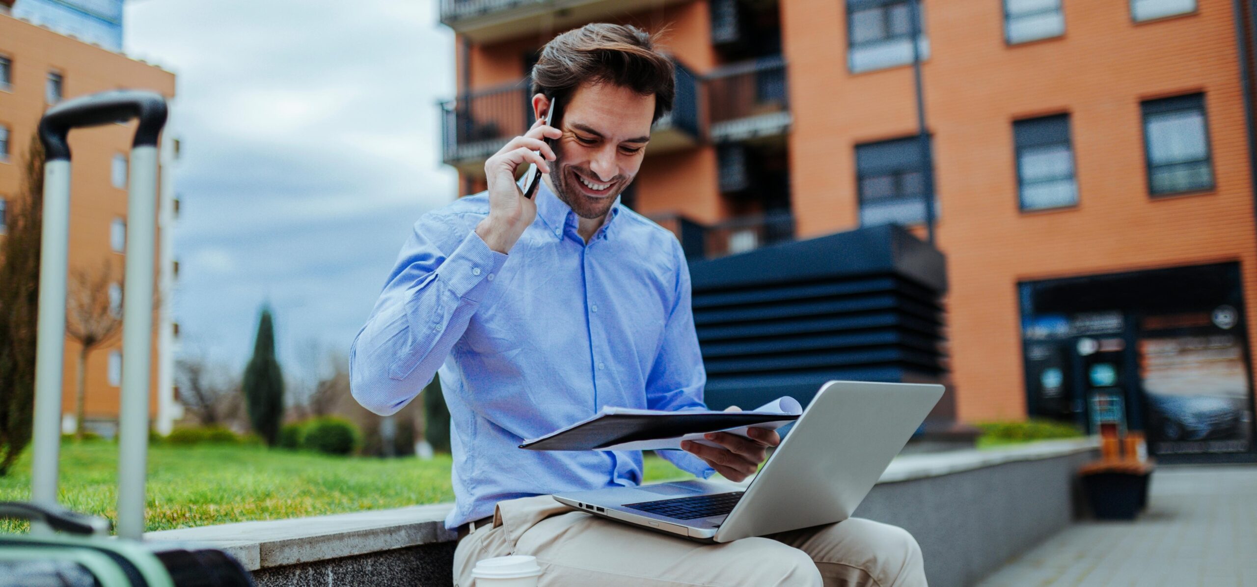 A businessman is on the phone with his colleague using a smartphone with a GOTAP smart tag behind him sitting on a garden chair in front of a large office wearing a blue shirt, beige trousers, laptop, and briefcase while holding a whiteboard with the business trip budget 2023.